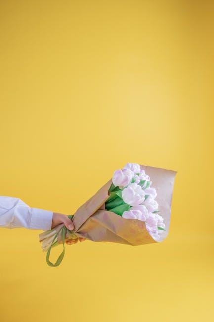 A person wearing a white sleeve holding a bouquet of fresh, pale pink tulips wrapped in light brown kraft paper and tied with a green ribbon, extending their arm against a vibrant yellow background. T