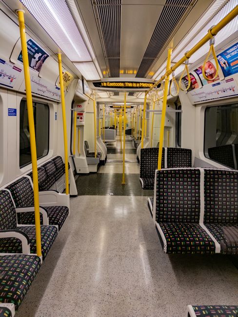 Empty interior of a modern metro train carriage with a clean, brightly lit environment featuring black seats with multicolored dotted patterns, yellow handrails and poles along the aisle, and large wi