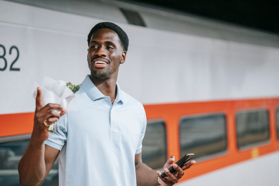 A smiling man with dark skin and short curly black hair is standing outdoors in front of a train with orange and gray exterior panels. He is holding a small bouquet of fresh flowers wrapped in white t