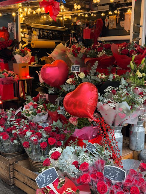 An outdoor floral display featuring a variety of fresh flowers including red roses, white baby's breath, and green foliage, arranged in baskets and bouquets. There are heart-shaped foil balloons in re