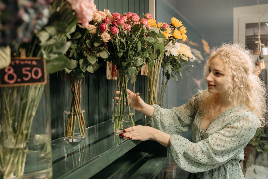 A young woman with curly blonde hair and light skin selecting fresh flowers at a floral shop, holding a glass vase with pink roses and yellow chrysanthemums. She is wearing a light-colored, long-sleev