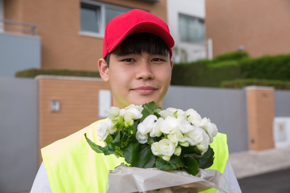 A young male flower delivery courier wearing a bright yellow high-visibility vest and a red cap, holding a bouquet of white roses wrapped in white paper. The roses have fresh green leaves, and the pet