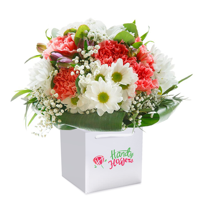 A striking, generously full bouquet is shown upright inside a neat white square gift bag, the composition revealing thoughtful floral design and textural contrast. The main blooms are sumptuous carnations in shades of soft coral-pink, warm peach and a rich, vibrant red; their frilled, layered petals form dense, velvety rosettes that radiate warmth. Nestled between these are numerous snowy white daisy-like chrysanthemums, each with a vivid yellow-green eye that provides crisp, joyful contrast to the carnation tones. Wisps of white baby's breath (Gypsophila) are woven throughout, introducing a feathery, cloud-soft texture that lightens the arrangement and invites a gentle inhalation of scent. A few closed buds with purple-tinged edges appear on the left-hand side, perhaps alstroemeria or similar, offering pockets of intrigue and a cooler tonality. The base is encircled by substantial, glossy deep green leaves that form a natural collar and lend architectural support to the rounded silhouette. The bouquet is placed in a premium white carrier bag with sturdy ribbon handles and photographed on a pristine white backdrop, the clean setting allowing every hue, petal edge and leaf sheen to register clearly. Under soft, even studio lighting the composition feels fresh, abundant and expertly finished - a ready-to-gift arrangement suited to anniversaries, congratulations or a heartfelt get-well visit within Ickenham and neighbouring Hillingdon streets.