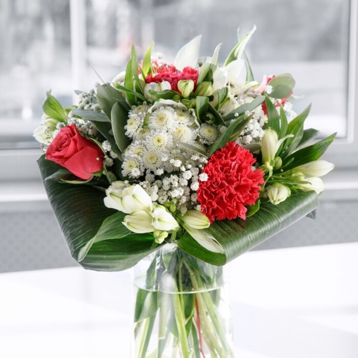 A carefully arranged bouquet in rich reds and fresh whites stands at the centre of this image, presented in a sleek, clear cylindrical glass vase that rests on a pristine white tabletop. The vase reveals a careful tangle of green stems beneath the waterline, trimmed and cleaned of excess leaves, reflecting the meticulous touch of an experienced florist. Above, the flowers fan out in a full, rounded form, with deep red roses drawing the gaze first. Their velvety petals fold and curl in perfect spirals, some just beginning to open, others a little more unfurled, giving the display a sense of movement and life. A large crimson carnation, heavily ruffled and richly textured, sits towards the front like a focal jewel, its frilled edges catching the light. Intermixed are pure white and creamy blooms: elegant white alstroemeria, each cluster presenting narrow petals with fine speckles near the throat, and several white lily buds still closed, their smooth, tapered shapes suggesting a quiet anticipation of blossoms yet to come. Tiny, cloud-like sprays of gypsophila, or baby's breath, drift through the arrangement, their small, starry white flowers softening the strong red-and-white contrast and lending the bouquet an ethereal, bridal touch. Around this core of colour, greenery provides structure and calm. Broad, glossy leaves are artistically looped and wrapped at the base, forming an attractive green collar, while slender, eucalyptus-style stems and pointed foliage reach upward at different angles, enriching the arrangement with layered shades of green. The background is softly lit and slightly blurred, reminiscent of a bright morning in an Ickenham home overlooking Swakeleys Park or a light-filled office near the station. The overall impression is of gentle sophistication and heartfelt warmth, suitable as a centrepiece for an anniversary dinner, a thank-you gift, or a thoughtful same-day delivery for someone special in the local area.