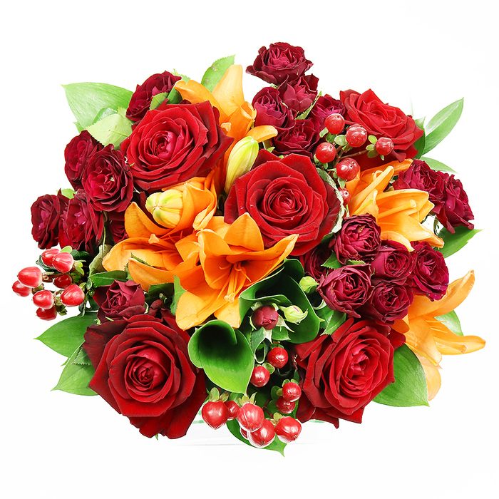 From a top-down viewpoint on a pristine white surface, this rich bouquet reads like a carefully staged still life, abundant with warm, celebratory colour and tactile contrast. The centre is anchored by several oversized deep red roses whose velvety petals unfurl in layered spirals, exuding a sense of classic romance and visual weight. Interleaved throughout are bright orange Asiatic lilies, their trumpet silhouettes and visible stamens introducing a lively geometry; some lilies are fully open and dramatic, others in elegant bud form, creating a feeling of unfolding time. Small clusters of dark burgundy spray roses sit close to the larger blooms, their tightly packed florets adding plush texture and a deeper, wine-toned accent. Here and there, clusters of shiny red hypericum berries add cheerfully glossy punctuation, reflecting the soft studio lighting like tiny beads of light. Lush green foliage-broad, glossy ruscus or salal leaves-lifts and separates the blooms, preventing visual heaviness and providing fresh, cooling contrast to the reds and oranges. The lighting is even and gentle, eliminating harsh shadows and highlighting petal texture, berry gloss, and leaf sheen. Imagined in a local context, this bouquet would feel at home on a mantel in an Ickenham cottage near Ickenham station or as a centrepiece for a small anniversary dinner in the village. It communicates warmth, celebration, and thoughtful craftsmanship: a tactile, scented composition meant to be held, inhaled, and remembered.