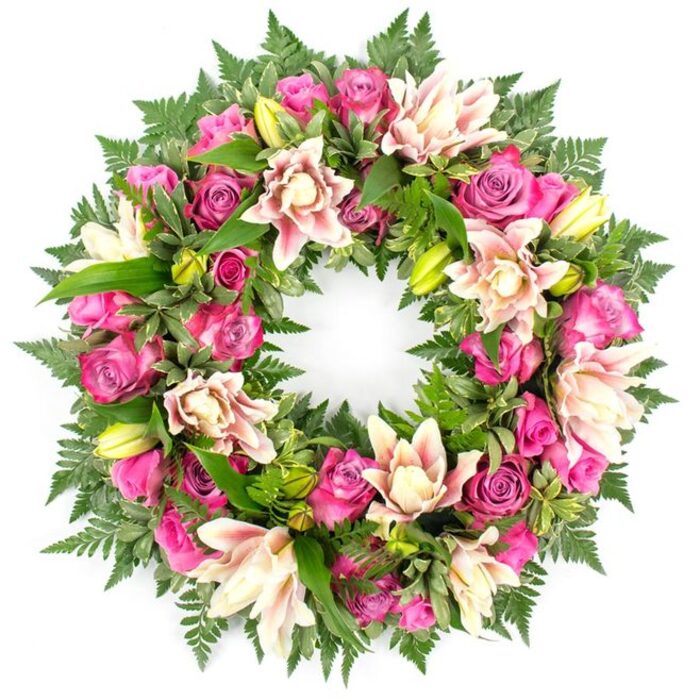 A richly detailed circular wreath fills the frame, photographed against a clean white background so that each flower and leaf can be appreciated. The wreath is constructed as a continuous ring, densely covered in florals that blend into one another, creating a seamless, full appearance often favoured for funeral and memorial tributes around Ickenham. The primary blooms are roses, their colours shifting from deep magenta and vibrant fuchsia to soft, powdery pink. Some roses are at the tight bud stage, with petals wrapped close to the centre, while others have opened wide, revealing multiple layers that create a lush, textured surface across the wreath. Among them are ample double lilies, their petals generously overlapping in tones of creamy white, light blush, and gentle peach. These lilies vary from fully open, showing delicate central stamens, to closed buds in fresh lime green that sit upright within the flowers, giving rhythm and height to the design. Lush greenery both supports and frames the blooms: delicate fern fronds extend outward around the edges, their feathery leaflets lending a soft fringe, while larger, smoother leaves in deep, glossy green fill gaps and add structure. Subtle touches of variegation in some foliage provide quiet visual interest. The white background removes any distraction from the composition, focusing attention on the interplay of pink petals against rich green foliage. The wreath carries a mood of tender dignity, a thoughtfully arranged floral expression that would rest beautifully beside a coffin in an Ickenham chapel, at a crematorium service, or in a family home during a private farewell gathering.