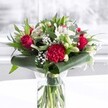 A rich, romantic bouquet in a clear cylindrical glass vase sits on a clean white surface, bathed in soft daylight like the kind that filters through bay windows along Ickenham High Road. Inside the vase, freshly cut green stems are visible below the waterline, neatly trimmed and aligned, giving a sense of care, craftsmanship, and just-delivered freshness. Above the glass, the arrangement swells into a generous dome of deep red and crisp white blooms, woven through with lush greenery. Velvety red roses are the stars of the piece, their petals spiralling open in perfect, classic rosettes that speak of passion and enduring love. Nestled among them, a large crimson carnation with finely ruffled, many-layered petals anchors the front of the display, its texture adding a touch of drama and movement. Threaded between the red blooms are white and cream flowers: graceful white alstroemeria with slender petals marked by delicate speckles, and several ivory lily buds held tightly closed, hinting at the promise of future blossoms. Tiny clouds of white gypsophila - baby's breath - are scattered throughout, creating an airy, almost mist-like softness that balances the bolder flowers. The foliage is varied and abundant, from broad, glossy dark green leaves that curve and fold around the base like a polished collar, to slimmer, eucalyptus-style sprigs and pointed leaves that add depth and contrast. The overall mood is uplifting, romantic, and quietly celebratory, ideal as a centrepiece for an anniversary dinner in an Ickenham home, a thoughtful Mother's Day surprise delivered to a favourite street near Swakeleys Park, or a refined gesture of affection for an office or reception area nearby. The composition feels both modern and timeless, as though crafted by a local florist with a particular love for elegant, heartfelt design.