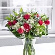 A vibrant mixed bouquet of red roses, red carnations, white alstroemeria, and clouds of white gypsophila stands proudly in a clear glass vase on a bright white tabletop, bathed in soft natural light from a nearby window in an Ickenham home. The deep crimson roses, some fully open with velvety spiralled petals and others still in tight buds, draw the eye first, their rich colour suggesting romance and heartfelt affection. Nestled closely beside them, ruffled red carnations add extra fullness and a soft, cushiony texture, echoing the same passionate shade while giving the bouquet a traditional, comforting feel. Between the stronger reds, clusters of white alstroemeria-some in bud, some just starting to open-introduce a creamy, trumpet-like softness, their pale petals tipped with a hint of green that harmonises with the foliage. Fine sprays of baby's breath are threaded throughout like tiny white stars, creating an airy, cloudlike veil that lightens the whole composition. Around the base, broad, glossy green leaves cradle the arrangement, while slimmer stems of lighter foliage weave upwards, adding gentle movement and depth. Through the clear vase you can see the neatly trimmed stems fanned out in clean water, reinforcing the sense of just-cut freshness you might expect from a local florist delivering across Ickenham and nearby Ruislip. The background is softly blurred and almost monochrome, with the outline of a large window and a muted outdoor scene, so the reds, whites, and greens appear even more vivid. The mood is one of cheerful appreciation and celebration-perfect for a birthday, a thank-you gift, or simply to bring a touch of warmth to a living room overlooking Swakeleys Park.