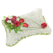 A solemn, hand-crafted sympathy pillow in soft rectangular form rests against a clean white background, its surface entirely covered in hundreds of small, pristine white chrysanthemums that give the piece a cloud-like, velvety texture. Around the edges a pale lime-green satin ribbon peeks through, gently framing the cushion and lending a subtle freshness to the overall composition. On the upper left corner a compact spray of deep red roses and rich burgundy carnations gathers, their velvety petals and ruffled edges forming a concentrated heart of color. Interspersed among them are little clusters of delicate white freesias or spray chrysanthemums that introduce brightness and delicate scent, while feathery fern fronds and glossy green leaves provide a soft, living backdrop. From this clustered spray a trio of slender, bright-green bear-grass arcs, like a calm brushstroke, sweep diagonally across the white chrysanthemum field toward the lower right, guiding the eye to a single, perfectly shaped red rose that rests there - a poignant focal point suggesting enduring love and remembrance. The arrangement is lit by gentle, diffuse light that highlights the texture of the petals and the soft sheen of the ribbon, and the overall impression is one of quiet dignity and comfort. As a florist who arranges memorial pieces for services in Ickenham - from small family churches to gatherings near Ickenham Common - I picture this pillow on a ceremony table or beside a photograph, its restrained palette of white, red, and green communicating respect, solace, and the subtle promise of peace.